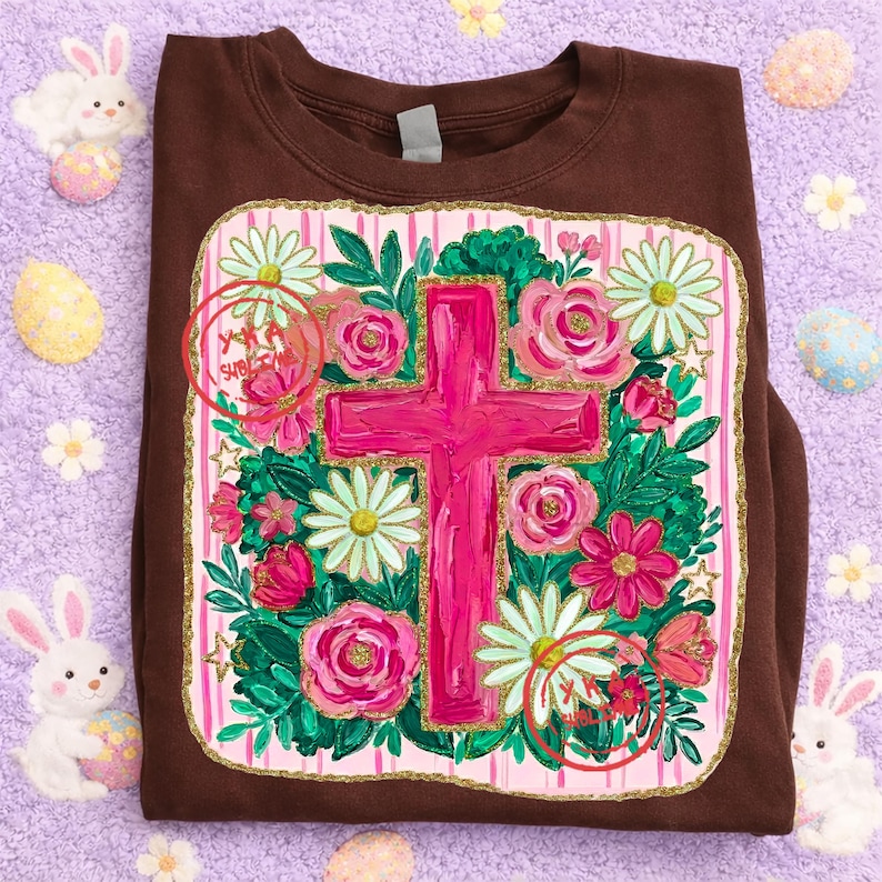 Peut inclure: Un sweat-shirt marron avec une croix rose entour&eacute;e de roses roses, de marguerites blanches et de feuilles vertes. Le motif est pos&eacute; sur un fond &agrave; rayures roses et blanches avec une bordure dor&eacute;e. Le sweat-shirt est pli&eacute; sur une surface violette.