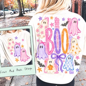 May include: A cream-colored sweatshirt featuring a graphic print of ghosts, stars, bats, and a pumpkin. The design includes the word BOO in large, colorful letters, a purple bow, and pink stripes. The print appears on both the front and back of the garment.