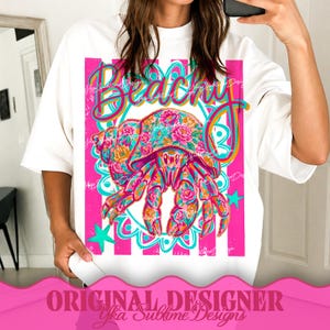 May include: White t-shirt featuring a vibrant graphic of a floral-patterned crab with the word "Beachy" in a cursive font. The design includes pink stripes, teal accents, and stars. The text "ORIGINAL DESIGNER Yka Sublime Designs" is at the bottom.