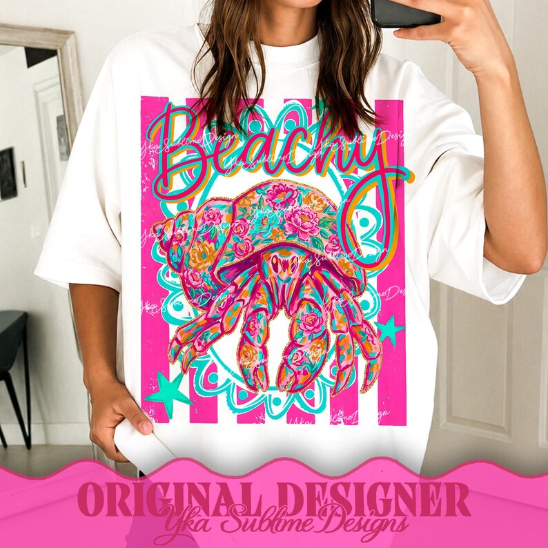 May include: White t-shirt featuring a vibrant graphic of a floral-patterned crab with the word "Beachy" in a cursive font. The design includes pink stripes, teal accents, and stars. The text "ORIGINAL DESIGNER Yka Sublime Designs" is at the bottom.