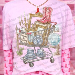May include: Pink t-shirt with a graphic of a shopping cart filled with vintage items, including cowboy boots, a disco ball, and blue and white china. The text "I'd Rather Be Thrifting" is on a sign. The shirt is worn over a pink long-sleeved shirt.