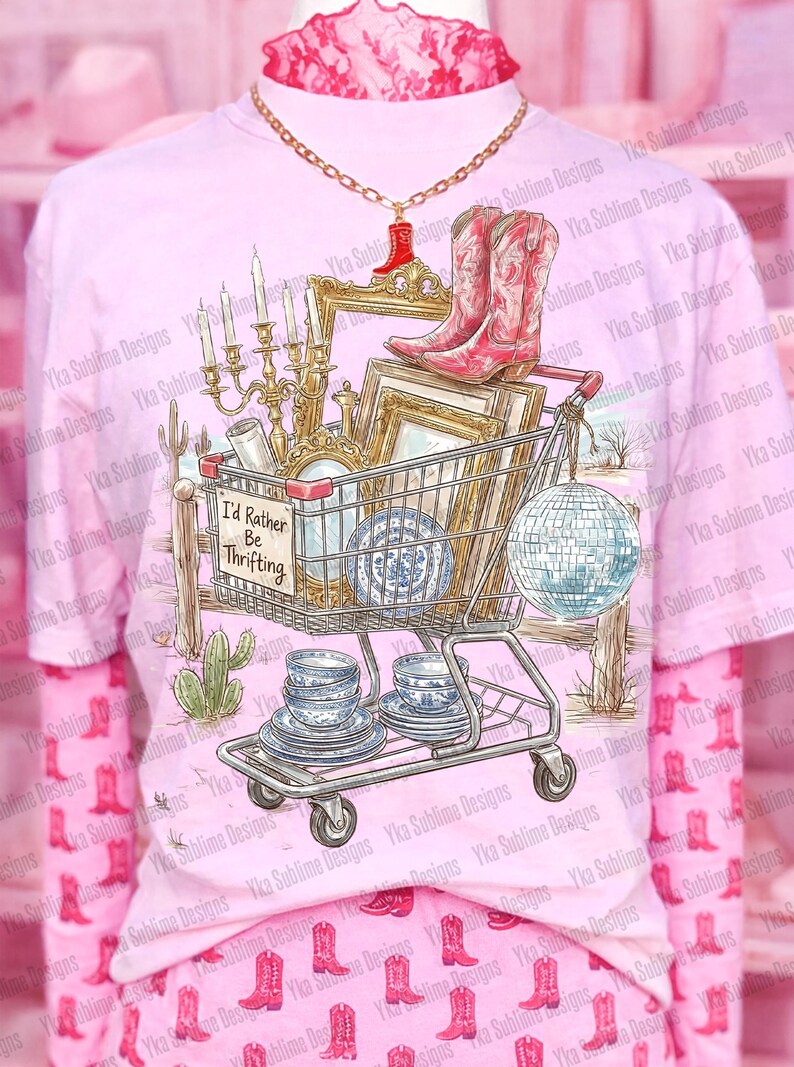 May include: Pink t-shirt with a graphic of a shopping cart filled with vintage items, including cowboy boots, a disco ball, and blue and white china. The text "I'd Rather Be Thrifting" is on a sign. The shirt is worn over a pink long-sleeved shirt.