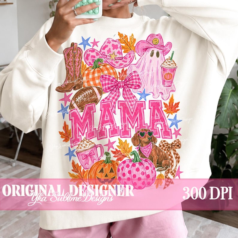 May include: A cream-colored sweatshirt featuring a colorful graphic print with the word MAMA in large pink letters. The design includes a pink cowboy hat, a ghost, a dachshund, pumpkins, a football, a bow, and autumn leaves in shades of pink and orange.