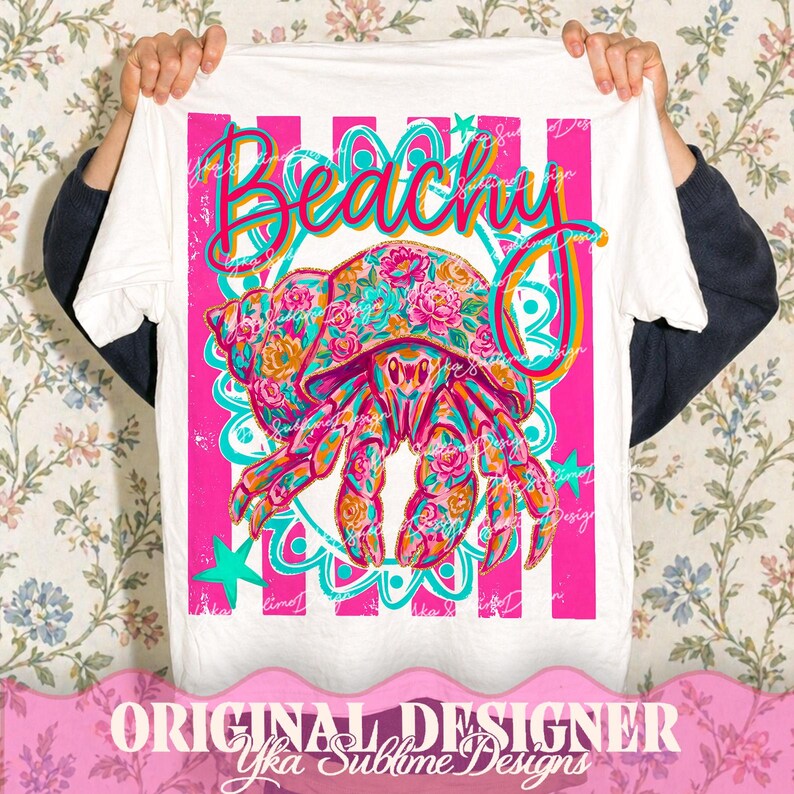 May include: White t-shirt with a vibrant graphic featuring a floral-patterned hermit crab and the word "Beachy" in a colorful script. The design includes pink stripes, teal accents, and stars, creating a summery vibe.