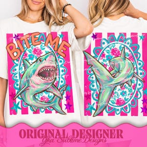 Può includere: T-shirt bianco con una grafica di squalo colorata e la scritta "BITE ME" in arancione. Il design include accenti rosa, turchese e blu, con un motivo floreale e a stelle. Un capo di abbigliamento casual.