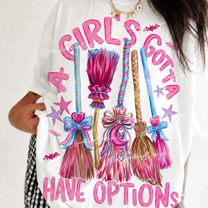May include: A white t-shirt featuring a graphic of four colorful broomsticks decorated with bows, hearts, and a crescent moon. The text "A GIRL'S GOTTA HAVE OPTIONS" is printed in pink letters around the brooms, accented by small purple stars and pink bats.