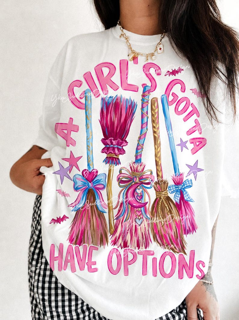 May include: A white t-shirt featuring a graphic of four colorful broomsticks decorated with bows, hearts, and a crescent moon. The text "A GIRL'S GOTTA HAVE OPTIONS" is printed in pink letters around the brooms, accented by small purple stars and pink bats.