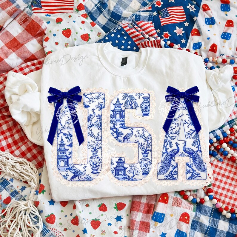 May include: White crew neck sweatshirt featuring the word "USA" in blue and white chinoiserie-style letters. Each letter is adorned with a blue velvet bow. The background includes red, white, and blue patriotic fabrics.