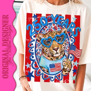 Op de afbeelding: Cr&egrave;mekleurig T-shirt met een luipaard met een blauwe hartvormige bril en een lichtblauw shirt met een Amerikaanse vlag. Het ontwerp bevat de tekst "250 YEARS" in blauw, met rode en blauwe sterren en strepen.