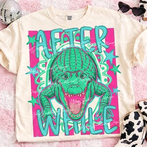 May include: Cream-colored t-shirt featuring a teal and pink graphic design. The design includes the words "AFTER WHILE" and a green alligator illustration. The shirt has a relaxed fit and short sleeves.