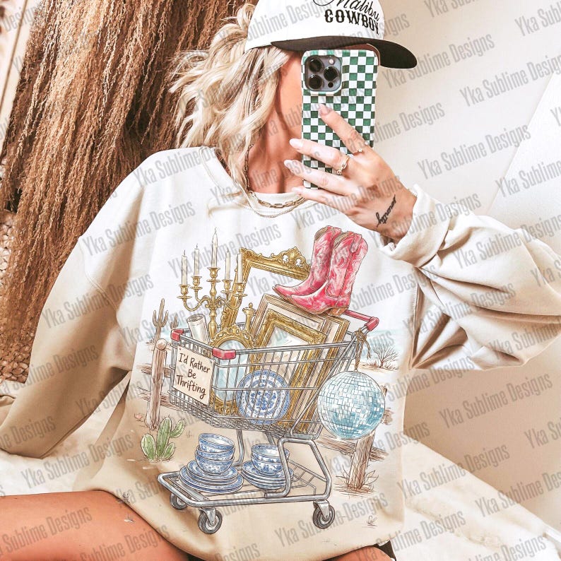 May include: Cream-colored sweatshirt with a graphic of a shopping cart filled with vintage items, including a candelabra, picture frames, and cowboy boots. The text "I'd Rather Be Thrifting" is on the cart. The design has a desert theme.