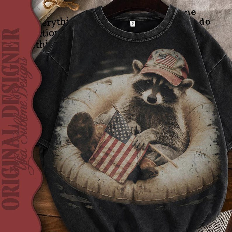 Puede incluir: Camiseta gris oscuro con un mapache que lleva una gorra de b&eacute;isbol con un dise&ntilde;o de la bandera estadounidense, descansando en una c&aacute;mara de aire y sosteniendo una peque&ntilde;a bandera estadounidense. La camiseta tiene un aspecto vintage y desgastado, con las palabras "ORIGINAL DESIGNER Yka Sublime Designs" impresas en el lateral.