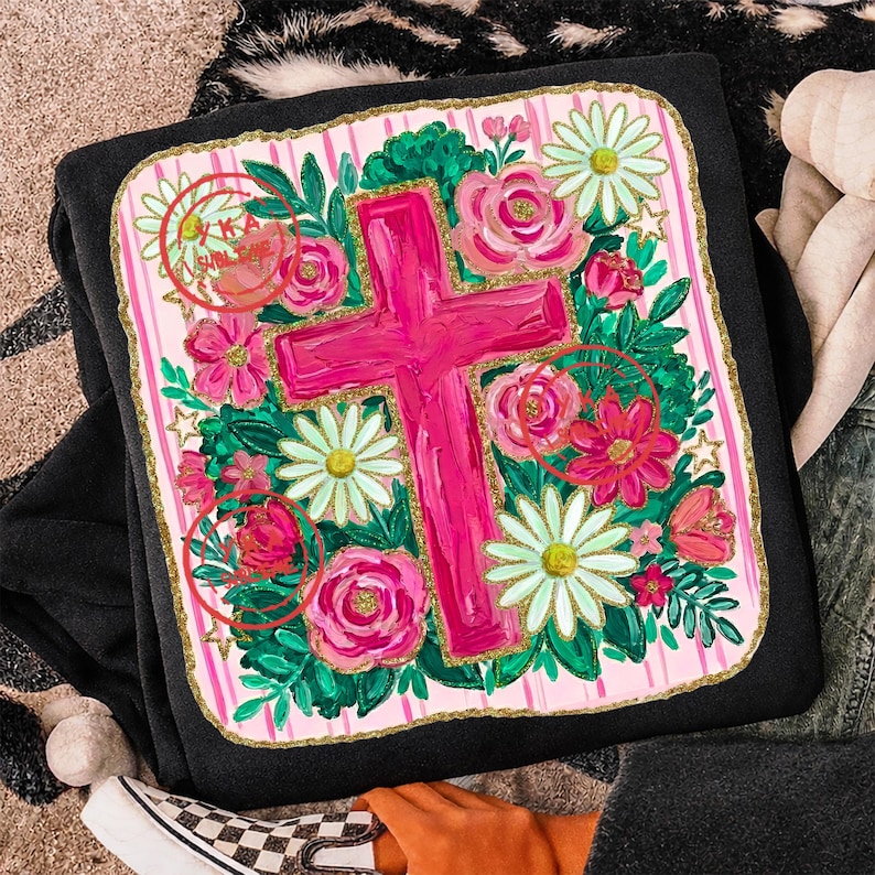 Peut inclure: Sweat-shirt noir orn&eacute; d'un motif floral vif avec une grande croix rose au centre. Le motif comprend des roses roses, des marguerites et du feuillage vert, le tout sur un fond &agrave; rayures roses et blanches avec une bordure dor&eacute;e.