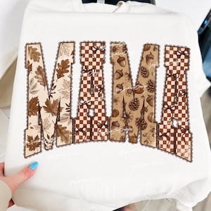 May include: A cream-colored crewneck sweatshirt featuring the word MAMA in large block letters. The letters are filled with a brown and white checkered pattern, autumn leaves, acorns, pinecones, and small bows, all outlined with a scalloped edge.