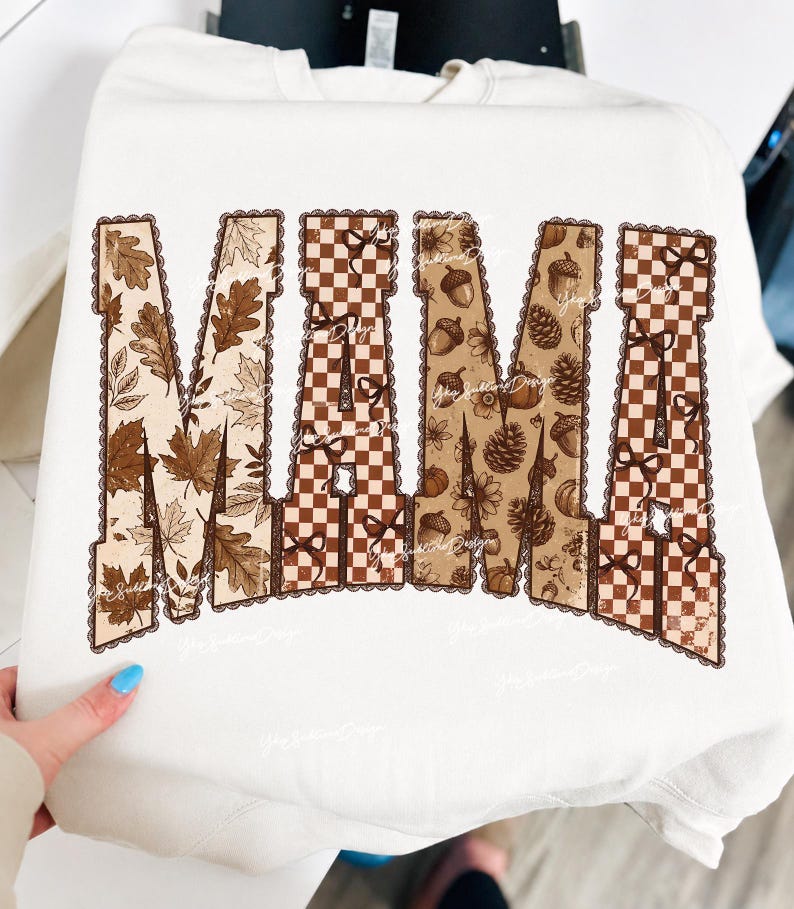May include: A cream-colored crewneck sweatshirt featuring the word MAMA in large block letters. The letters are filled with a brown and white checkered pattern, autumn leaves, acorns, pinecones, and small bows, all outlined with a scalloped edge.