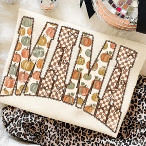 Puede incluir: Una sudadera de color crema con la palabra MAMA en letras grandes. Las letras están rellenas con un patrón de varias calabazas y un diseño de cuadros marrones y blancos, acentuado con pequeños lazos marrones y bordes festoneados.