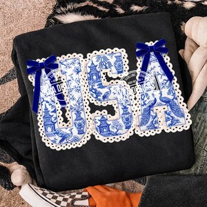 May include: Black sweatshirt featuring the word "USA" in a blue and white porcelain design. The letters are trimmed with white lace and decorated with blue velvet bows. The design includes pagodas, vases, and birds.