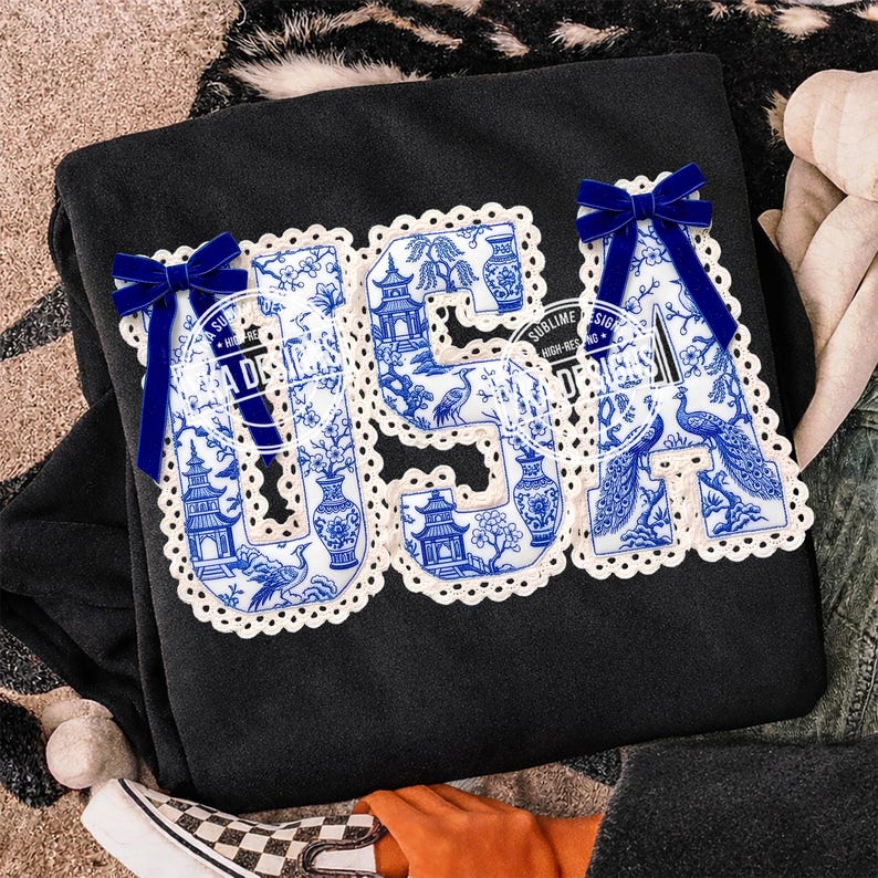 May include: Black sweatshirt featuring the word "USA" in a blue and white porcelain design. The letters are trimmed with white lace and decorated with blue velvet bows. The design includes pagodas, vases, and birds.
