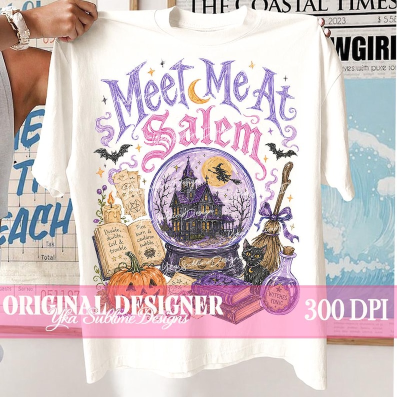 May include: A cream-coloured t-shirt featuring a graphic print with the text 'Meet Me At Salem'. The design includes a purple crystal ball with a haunted house, a witch on a broom, a black cat, a pumpkin, an open spellbook, and a potion bottle.