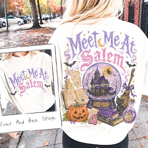 May include: An off-white t-shirt featuring a graphic design with the text Meet Me At Salem. The illustration includes a purple crystal ball with a haunted house, a black cat, a broom, a pumpkin, an open book, and a bottle labelled Witches Tonic.