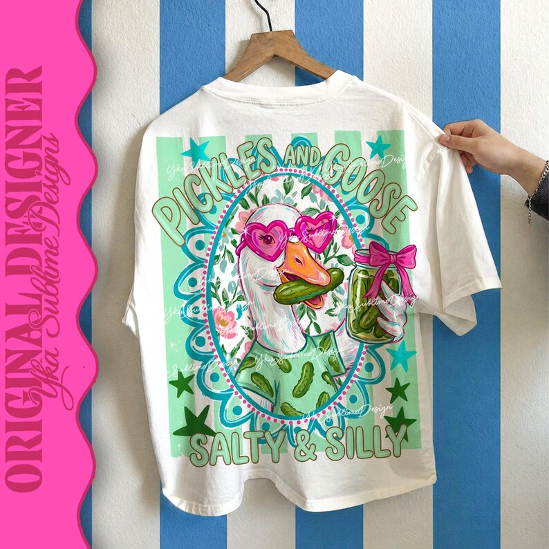 May include: White t-shirt with a whimsical graphic featuring a goose wearing heart-shaped sunglasses and holding a pickle. The design includes the text "Pickles and Goose" and "Salty & Silly" in a playful font. The shirt is hanging on a wooden hanger.
