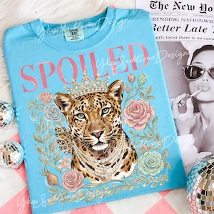Puede incluir: Camiseta azul claro con la palabra "SPOILED" en rosa. La camiseta presenta un leopardo con corona y collar de perlas, rodeado de rosas y hojas. La camiseta es de "Yka Sublime Design".
