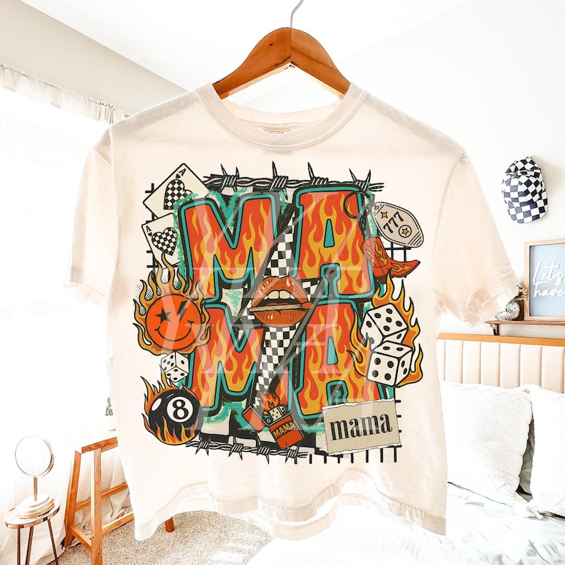 Puede incluir: Camiseta color crema con un dise&ntilde;o gr&aacute;fico vibrante. El dise&ntilde;o presenta la palabra "MAMA" en letras audaces en forma de llama, junto con dados, naipes, una carita sonriente y otros elementos retro. La camiseta est&aacute; colgada en una percha de madera.