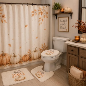 May include: A bathroom set featuring a shower curtain, toilet lid cover, and two floor mats, all decorated with a cream background, orange autumn leaves, and pumpkins. The mats and lid cover display the text Hello Fall in a brown script font.