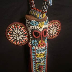 May include: A beaded mask with a dark background. The mask features a central face with red, black, and white details, flanked by circular ear-like elements. The mask has a long, beaded body with blue, yellow, and red accents.