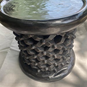 May include: A dark gray, hand-carved wooden stool with a round, shallow top and a textured, geometric patterned base. The stool is made of a dark wood and has a smooth, polished finish. The overall design is simple yet elegant.