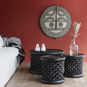 May include: A living room with three dark wooden side tables with carved lattice patterns. A large, circular tribal mask is on the red wall. Two white vases and a clear vase with a plant are on the tables.