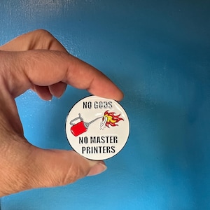 May include: A round, white button with the text "NO GODS NO MASTER PRINTERS" in black. The button features a red and silver spray can with flames. The button is held in a hand against a blue background.