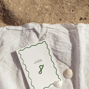 May include: A white table number card with a green wavy border and the word "Table" and the number "8" in green. The card rests on a white cloth with two seashells, with sand in the background.