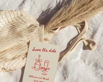 Może przedstawiać: Kremowe zaproszenie ślubne z czerwonym tekstem "Save the date" oraz imionami "Catherine and Daniel" i datą "11.12.2030". Zaproszenie leży obok beżowej siatki, okularów przeciwsłonecznych i kępy suchej trawy.