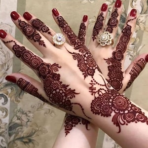 May include: Two hands with intricate henna designs in a deep burgundy color. The henna patterns feature floral and swirl motifs. The nails are painted a matching burgundy. Two ornate rings with sparkling stones adorn the fingers. The text "@hennabysana809" is visible.