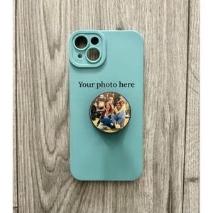 May include: A teal phone case with a circular photo grip showing a family portrait. The case has a camera cutout and the text "Your photo here" printed on the front. The background is a wooden surface.