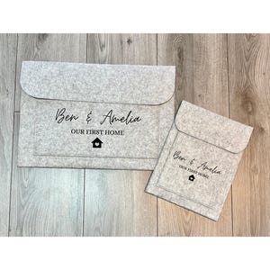 Può includere: Due portadocumenti in feltro grigio. Quello più grande ha il testo "Ben & Amelia OUR FIRST HOME" con un'icona a forma di casa. Quello più piccolo ha lo stesso testo e icona. Entrambi hanno una chiusura a patta.
