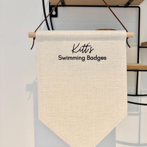Pode incluir: Bandeira de tecido creme com o texto "Kitt's Swimming Badges" em escrita preta. A bandeira é suspensa por um pino de madeira com correias de couro marrom. A bandeira tem um fundo pontiagudo.