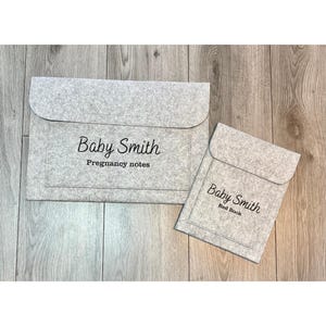 以下が含まれることがあります： 丸みを帯びたフラップが付いた2つの灰色のフェルトドキュメントホルダー。大きい方には黒い筆記体で「Baby Smith Pregnancy notes」と書かれています。小さい方には「Baby Smith Red Book」と書かれています。どちらも木目調の表面にあります。