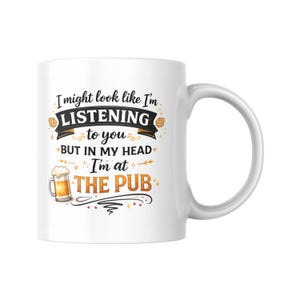Puede incluir: Taza de cerámica blanca con asa curva. La taza presenta el texto "I might look like I'm LISTENING to you BUT IN MY HEAD I'm at THE PUB" en letras negras, doradas y naranjas, con un gráfico de cerveza.