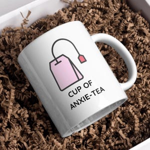 May include: White ceramic mug with a handle, featuring a pink tea bag graphic and the text "CUP OF ANXIE-TEA" in black. The mug is presented in a white box filled with brown crinkle paper.
