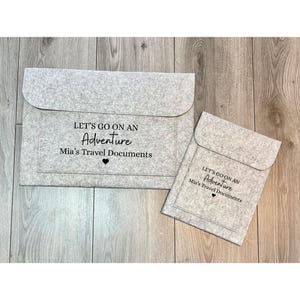 Peut inclure: Deux pochettes de documents en feutre gris. La plus grande est rectangulaire avec une fermeture à rabat. La plus petite est de style enveloppe. Les deux portent l'inscription "LET'S GO ON AN Adventure Mia's Travel Documents".