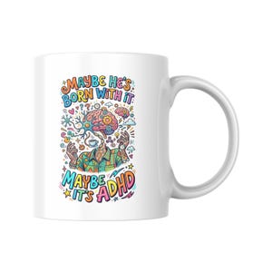 May include: White ceramic mug with a colourful illustration. The design features a brain with gears, a coffee cup, and the text "MAYBE HE'S BORN WITH IT, MAYBE IT'S ADHD" in a playful font. The mug has a curved handle.