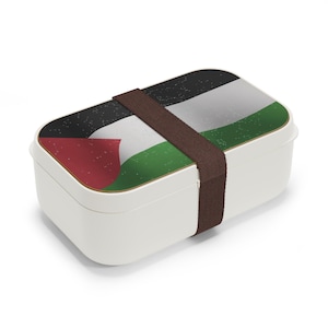May include: A white rectangular lunch box with a brown strap. The lid displays the flag of Palestine, with red, white, green, and black colours. The lunch box is designed for carrying food.