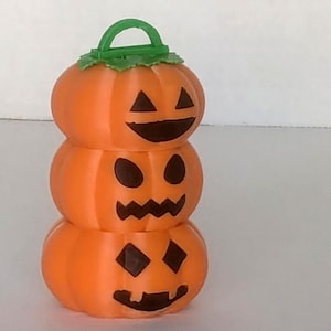 May include: A stacked orange pumpkin decoration with three sections, each featuring a carved jack-o'-lantern face. The top pumpkin has a green handle and leaf detail. The faces are black, and the overall design is festive.