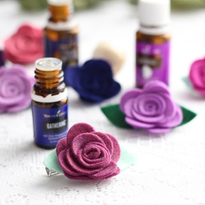 Pode incluir: Uma variedade de presilhas de cabelo com flores de feltro coloridas e frascos de óleos essenciais. As presilhas de cabelo são em tons de roxo, rosa e bordô, com detalhes de folhas verdes. Os frascos de óleos essenciais têm rótulos azuis e roxos.