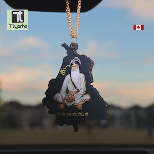 May include: A black car charm featuring an image of a religious figure, shaped like a map. The charm is suspended by a gold chain. The image includes text in a foreign language. The Tlysta logo is in the upper left corner.