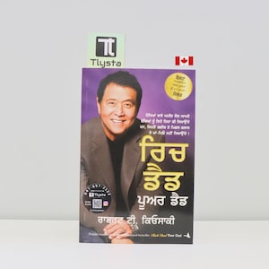 May include: A book titled "Rich Dad Poor Dad" in Punjabi, featuring a portrait of a smiling man on the cover. The book has a purple background with text and a gold seal. A Canadian flag is visible.