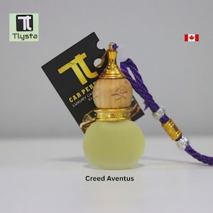 May include: A small, round, light green perfume bottle with a wooden top and gold accents. The bottle is attached to a purple cord. A black tag with gold text reads "CAR PERFUME LUXURY CAR EL". The text "Creed Aventus" is below the bottle.