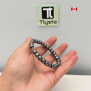 May include: A silver beaded bracelet with a clasp, held in a hand. The bracelet is made of small, round beads. The background includes a sign that says "Tlysta".